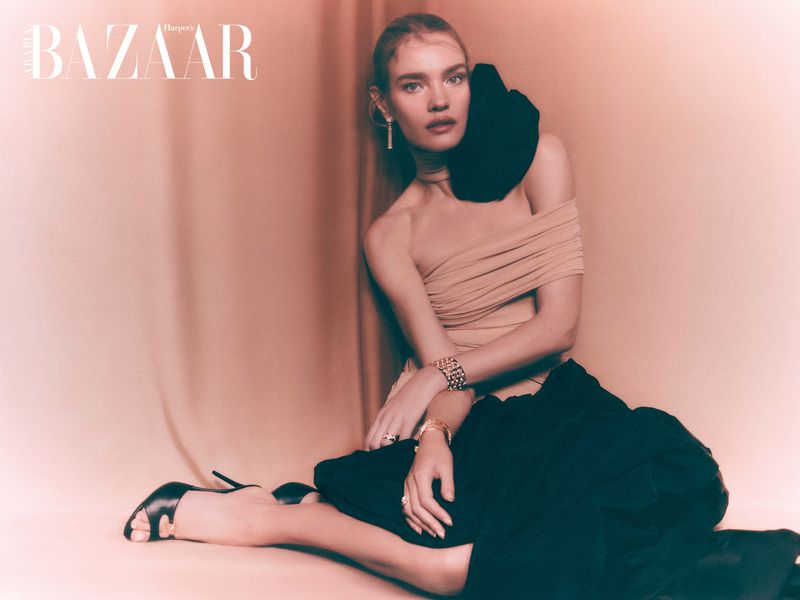 Editorial fashion shot of a woman in beige top and black skirt posing on a peach-colored surface in a studio setting, elegant and graceful.