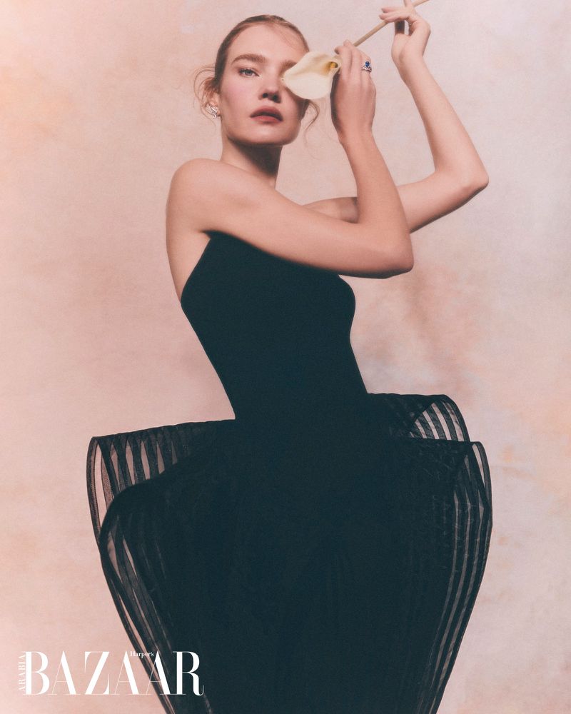 Portrait of a fair-skinned woman in a black dress holding a white flower, set against a soft pink backdrop in a fashion editorial.