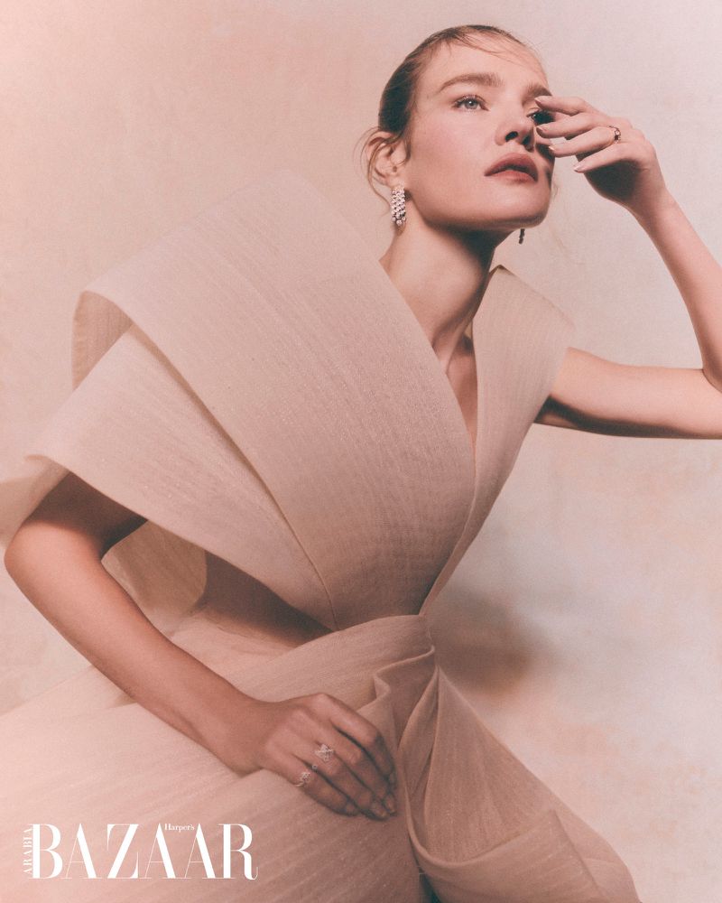 Elegant woman in a voluminous beige dress and jewelry poses in a studio with diffused lighting for a fashion editorial shot.