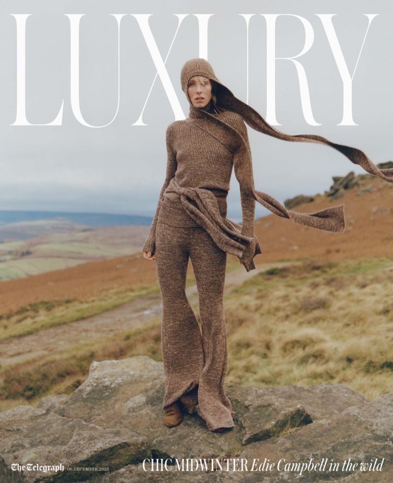 Fashion editorial shot of a model in a brown knit outfit posing on a rock outcrop overlooking a hilly landscape with a cloudy sky.