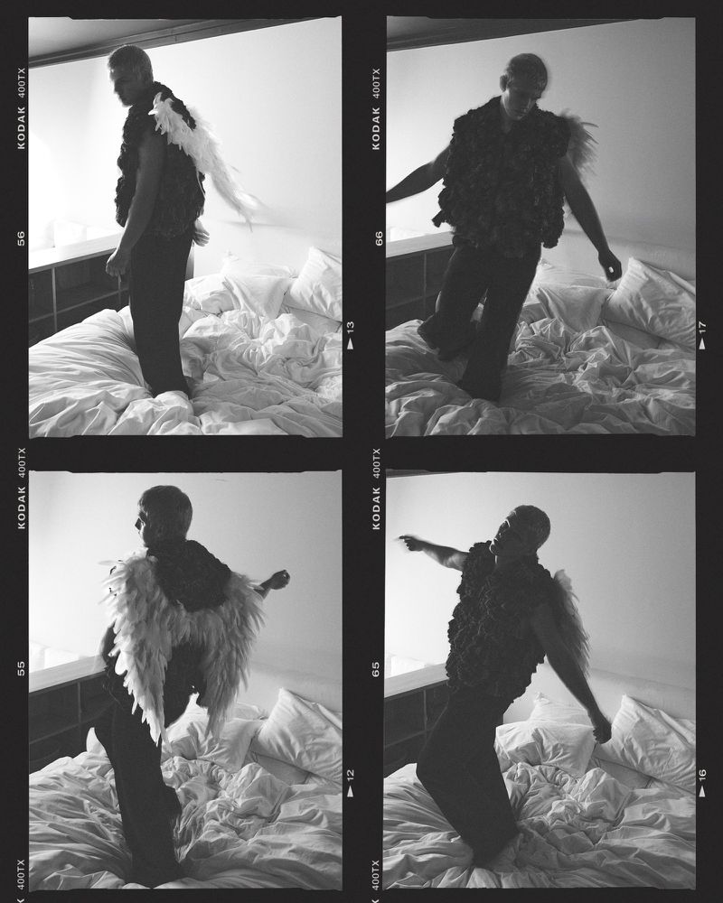 Monochrome photo of a man posing on a bed in a vest with wing-like extensions, captured in a film strip style image with grain.