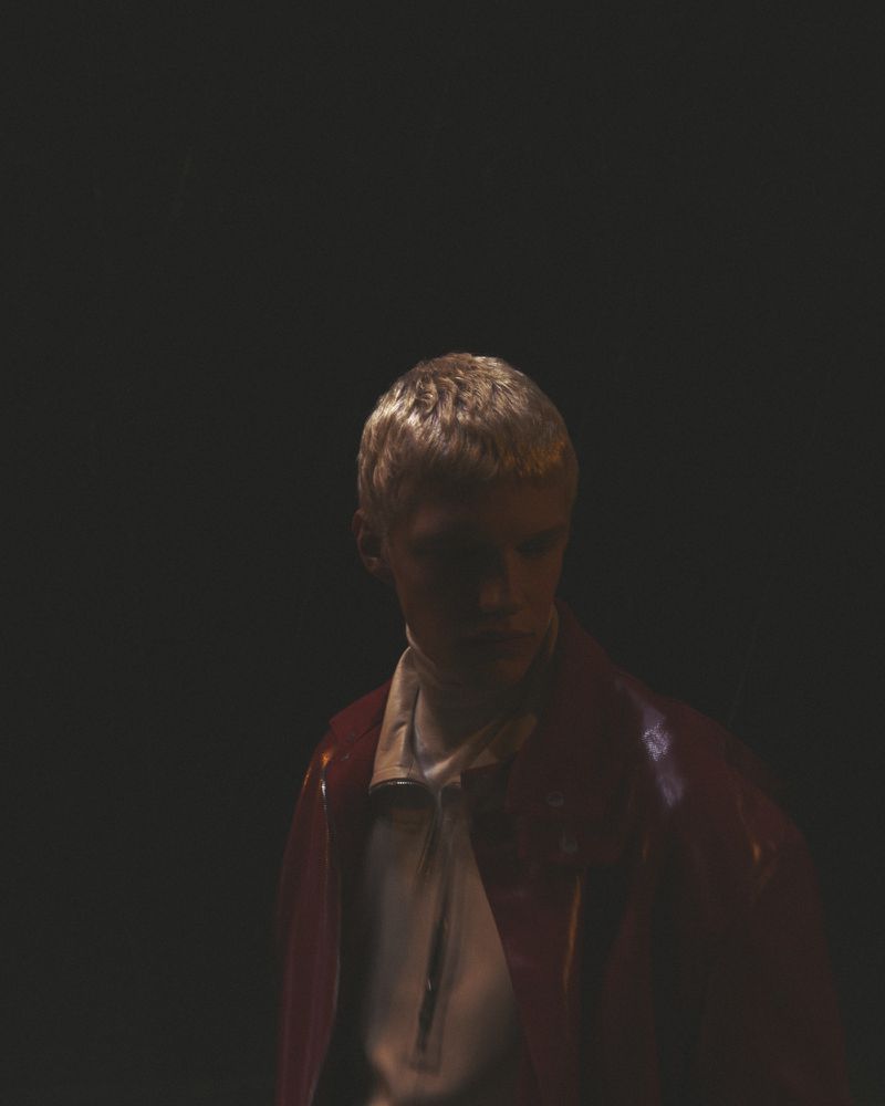 A young adult poses in a portrait. The person wears a white shirt and red jacket against a dark background. Studio lighting is used.