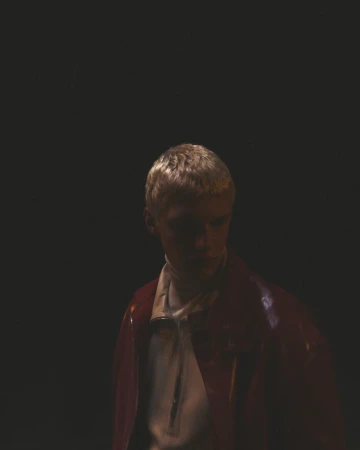 A young adult poses in a portrait. The person wears a white shirt and red jacket against a dark background. Studio lighting is used.