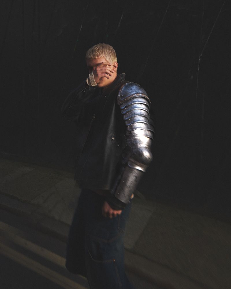 Adult male poses with armor and covers face partially with hand in dark setting with hard lighting emphasizing metallic textures.