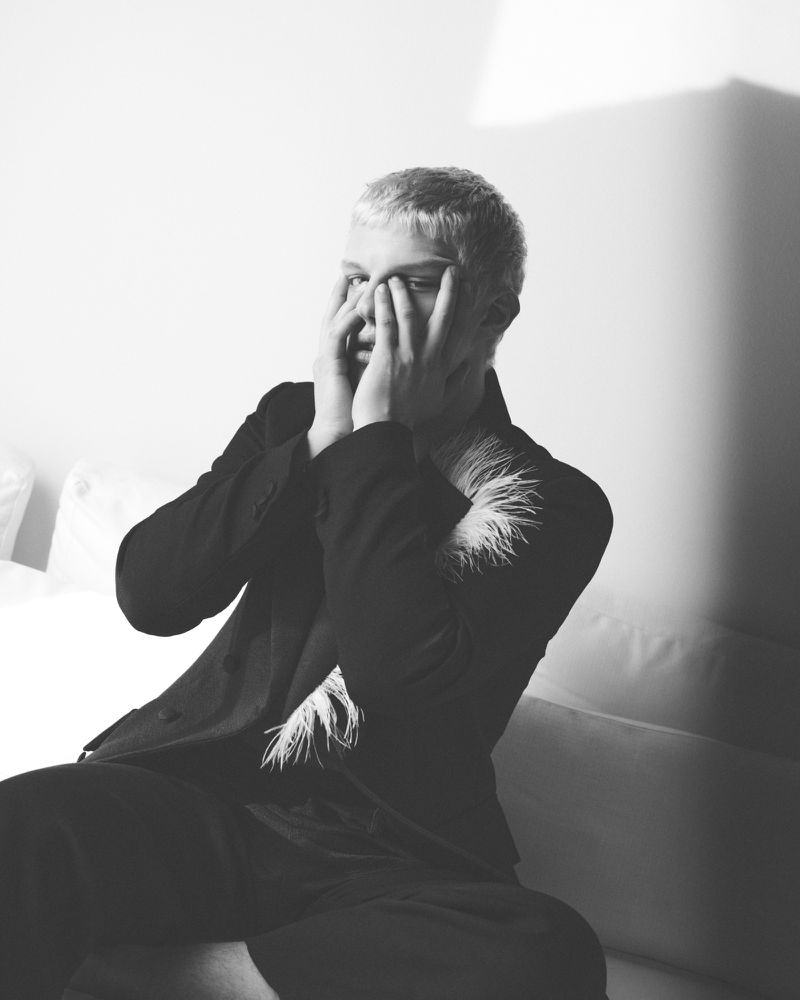 Monochrome portrait of an adult with hands covering face, wearing dark suit with feather detail, against a light wall with shadows.