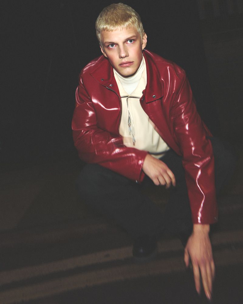 Young adult male in a red leather jacket crouching in a dimly lit indoor environment, looking away from the camera.
