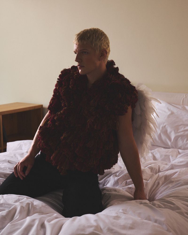 Adult with blonde hair kneels on a bed, facing away from the camera, wearing a maroon garment and feathery wings against a neutral wall.