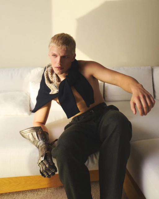 Blond male model with a metal gauntlet poses on a white couch in a studio setting with a dark sweater draped over his shoulders.
