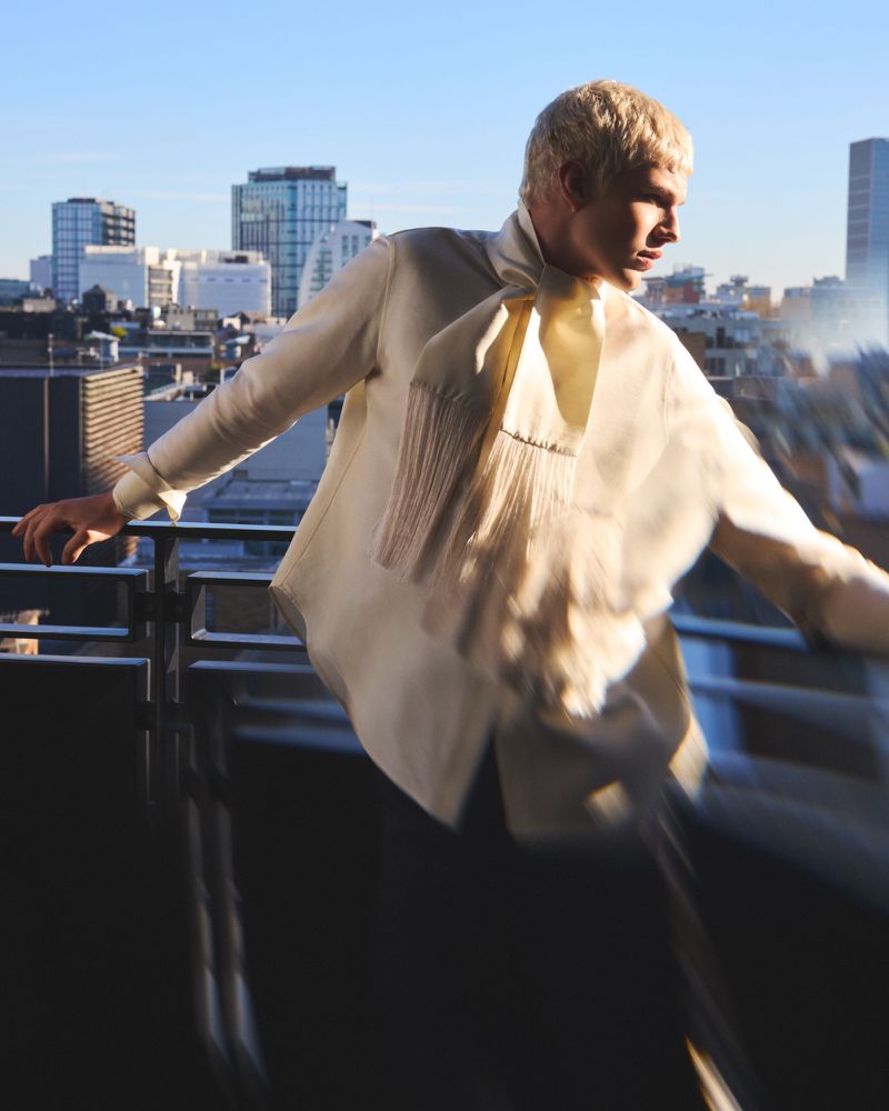 Man in a light-colored shirt with a scarf-like detail stands on a city balcony with a cityscape background under a clear blue sky.