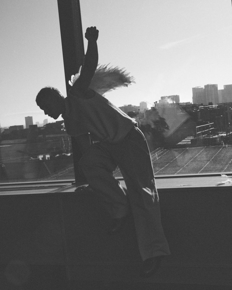 Black and white photo of a person with wings silhouetted against an urban skyline, arms raised in a triumphant pose, captured indoors.