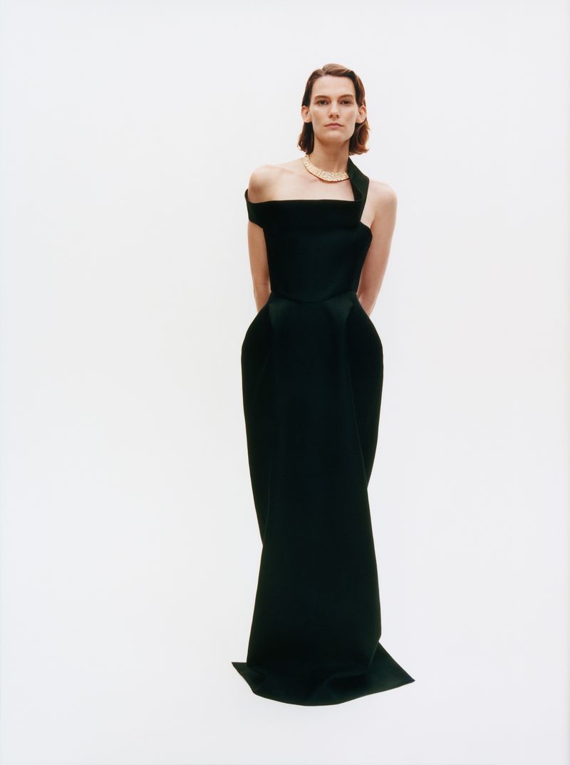 Elegant woman in a full-length black gown poses in a white studio. A gold necklace adorns her neck, and her brown hair is neatly styled.
