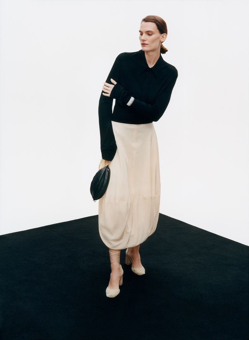 A woman in a black sweater and light skirt poses on a black and white floor, holding a black purse and wearing beige heels.