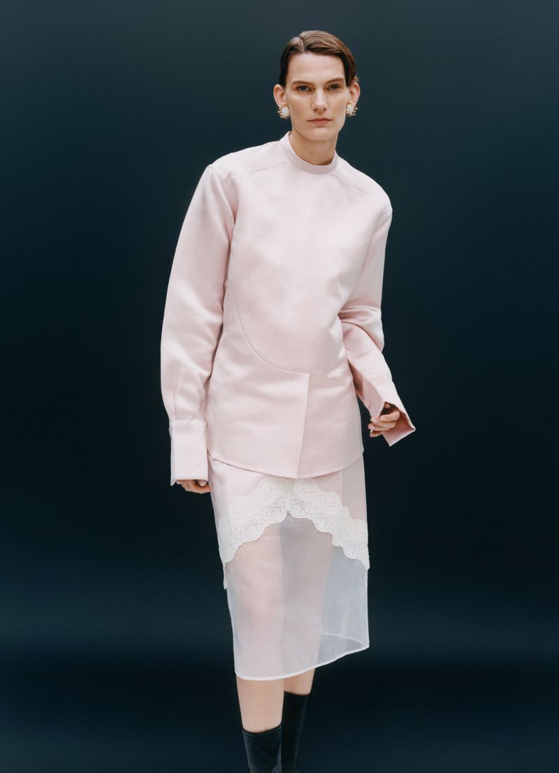 Fashion model poses in a pale-pink jacket and sheer white skirt against a dark background. The image features a fashion-forward style.
