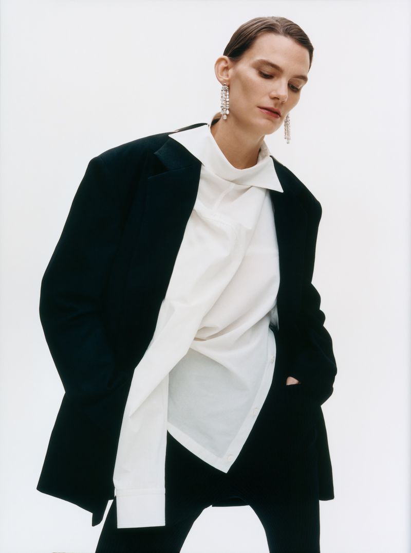 Model Lena Hardt wearing an oversized black blazer, draped white shirt, and dangling earrings against a white seamless background.