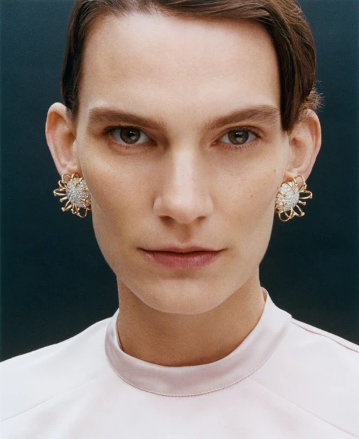 Close-up of a model wearing flower-shaped diamond earrings. The model has short brown hair, brown eyes, and a light pink top.