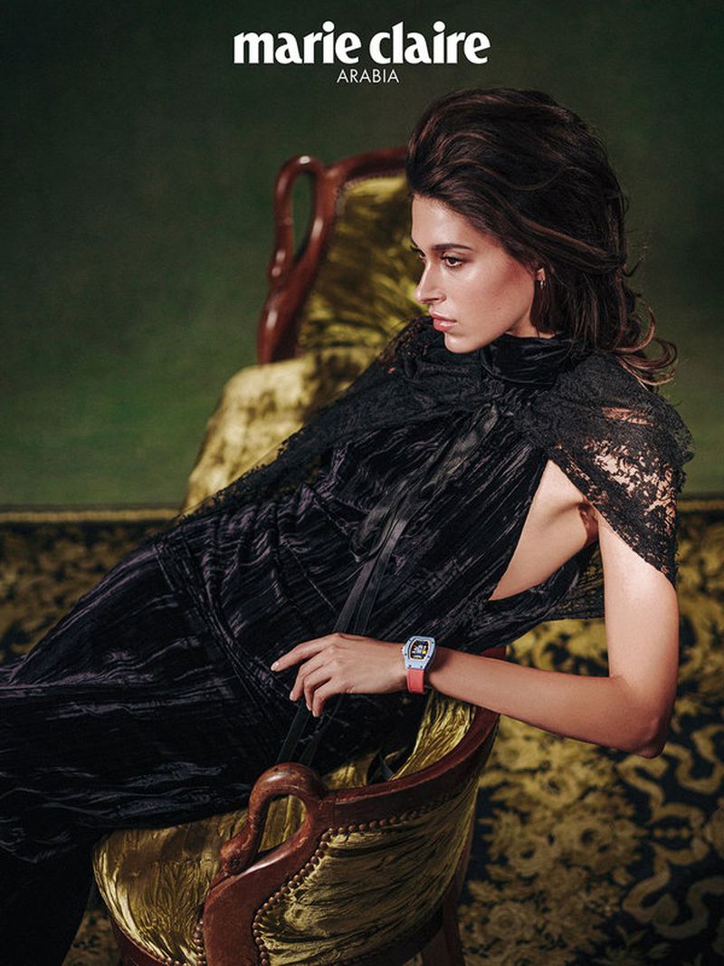 Fashion editorial shot of a woman reclining on a chair, wearing a black lace dress and a wristwatch, with a dark green background.