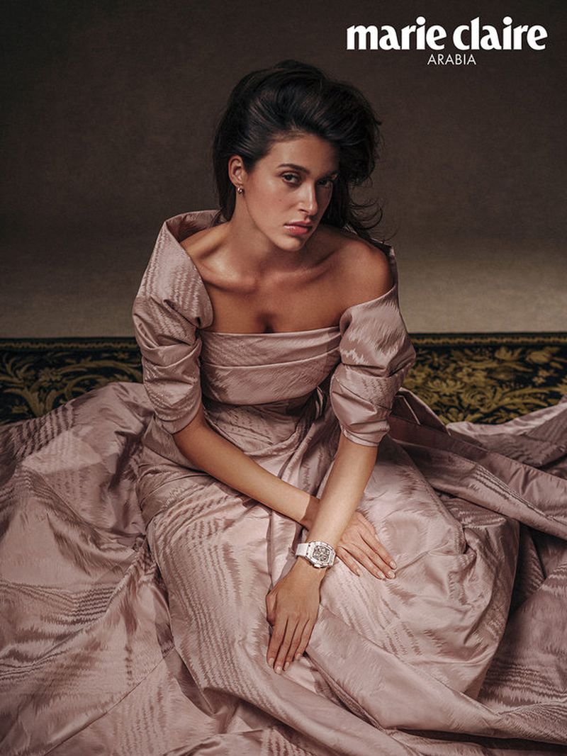 Elegant woman in flowing pink off-shoulder gown poses on patterned rug in a close-up fashion shot for Marie Claire Arabia magazine.