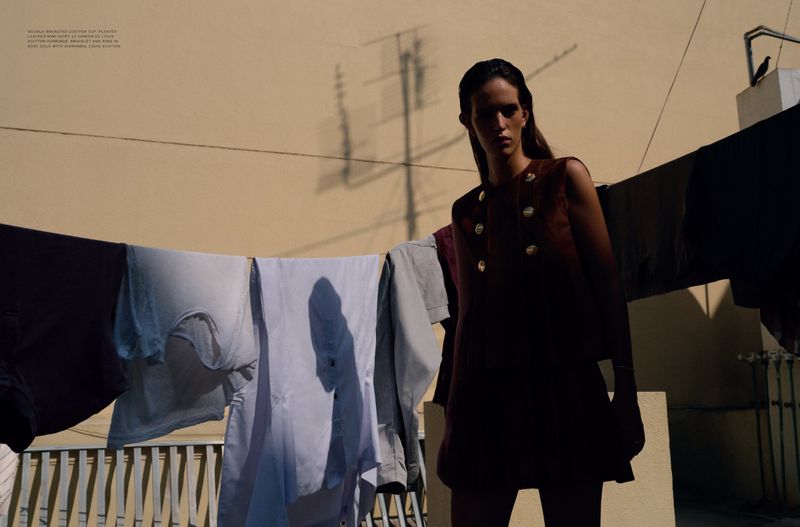 A model poses in a brown two-piece outfit with gold buttons on an outdoor rooftop with clothes hanging on a line in soft sunlight.