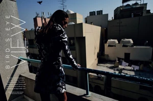 Model Anica Cuca in a black and white patterned dress leaning on a blue balcony railing on a sunny city rooftop.