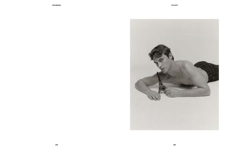 A shirtless adult male model poses while laying on a white surface and holding a metal Eiffel Tower miniature replica in a studio shot.