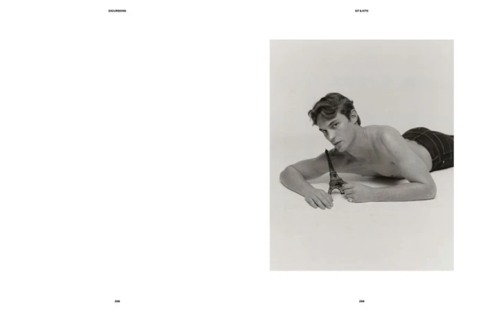 A shirtless adult male model poses while laying on a white surface and holding a metal Eiffel Tower miniature replica in a studio shot.