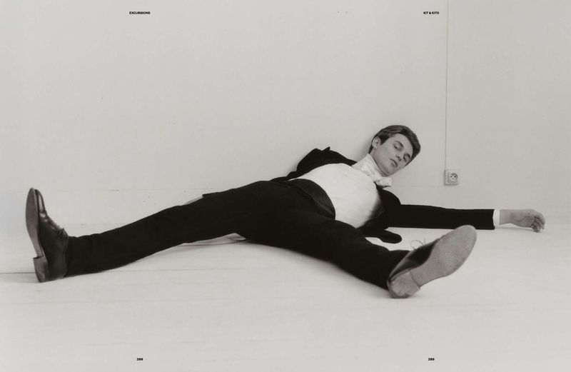 Man in a suit lies on the floor with arms and legs outstretched against a white wall, his head turned to the side in a fashion pose.