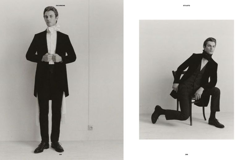 Two men in formal black suits pose in a studio. One stands, the other sits on a chair. High contrast monochrome studio fashion shot.