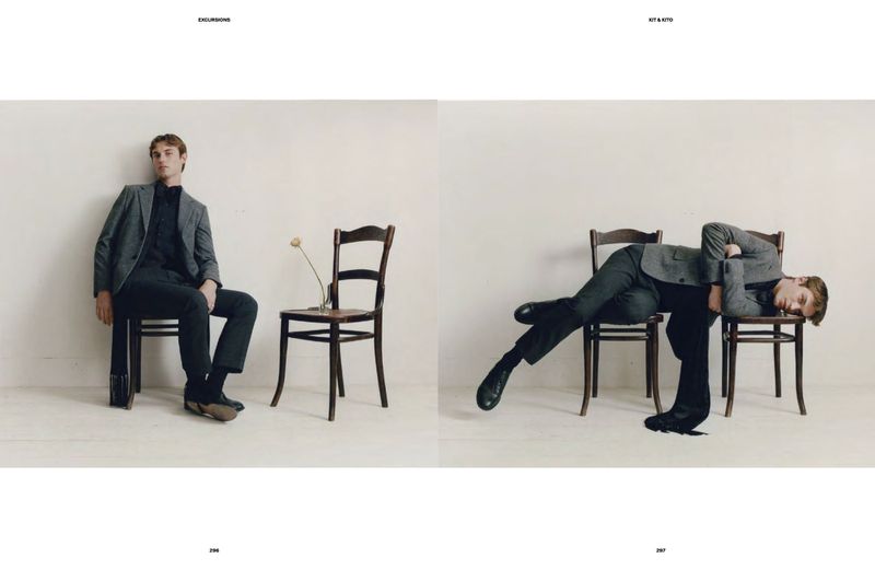 Editorial fashion shot of a man in a grey suit sitting and laying on a wooden chair against a neutral wall background with soft lighting.