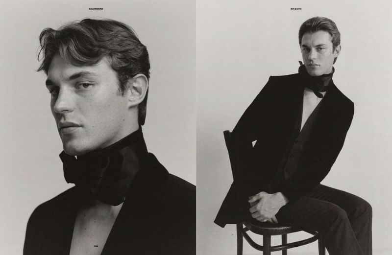 Black and white fashion shot of a male model in a blazer with a large neck bow, sitting on a stool against a white backdrop.