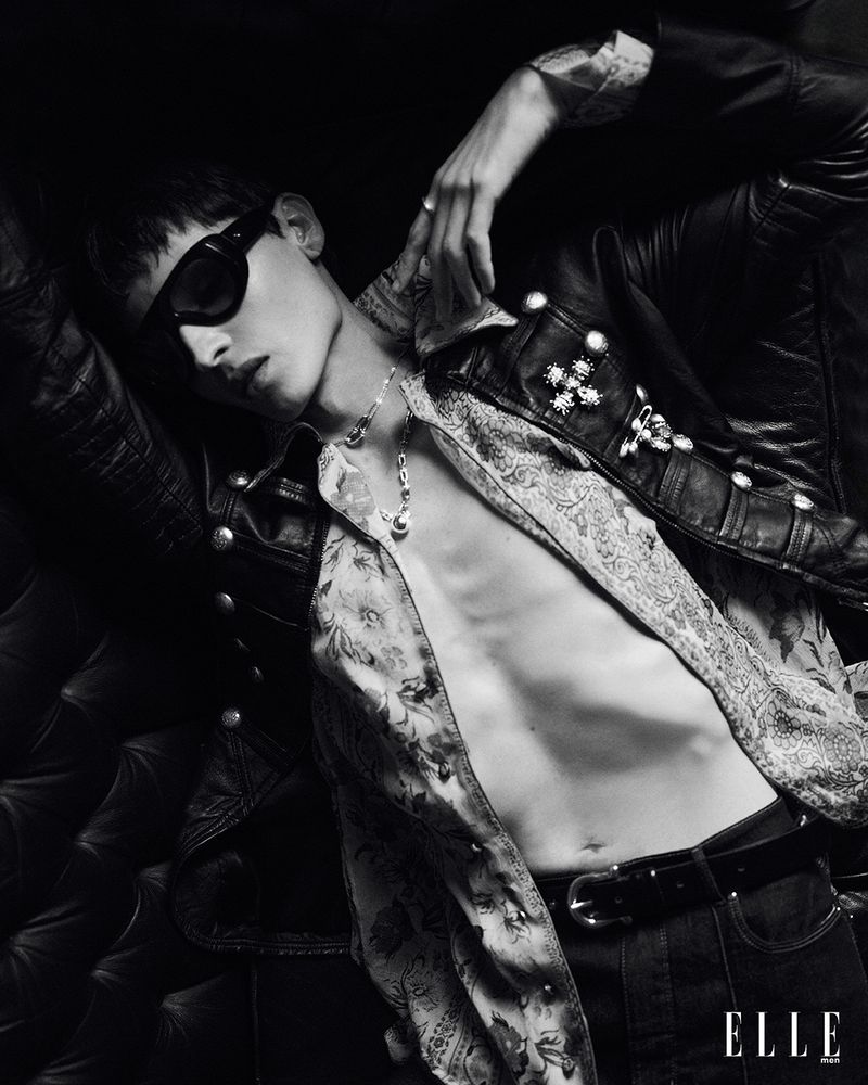 Black and white studio fashion shot of a young adult man in a leather jacket and unbuttoned shirt reclining on a black leather couch.