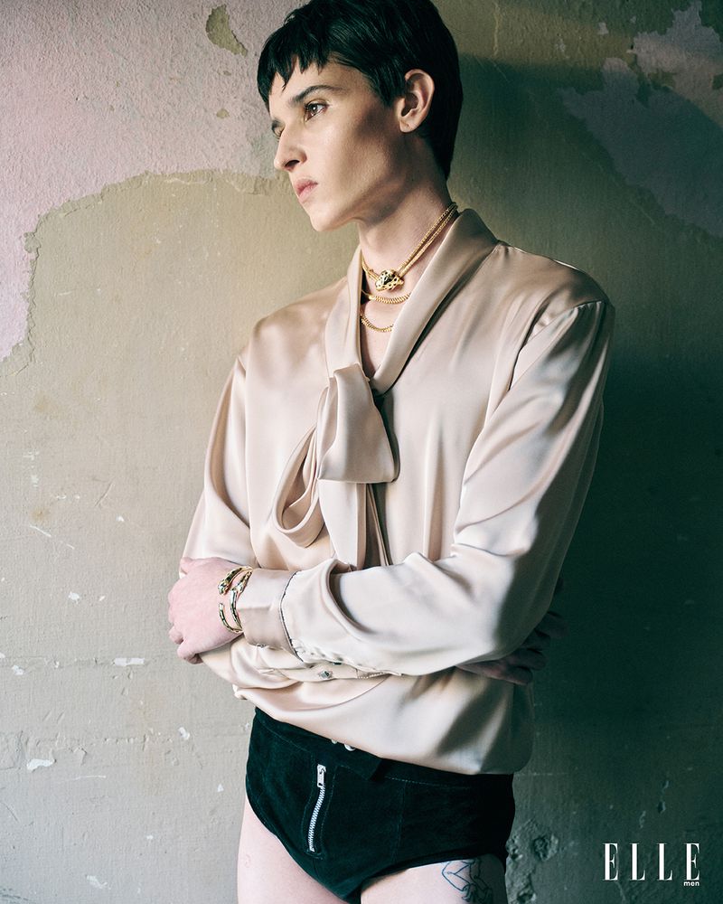 Portrait of a slender adult with crossed arms in a champagne-colored blouse, black shorts, and jewelry, posing against a textured wall.