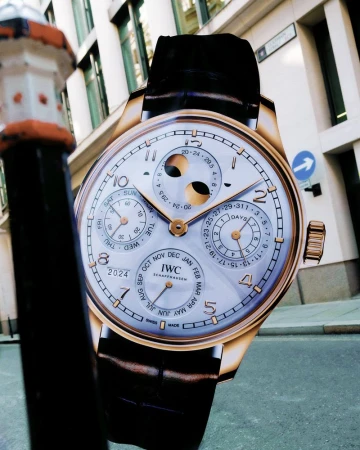 Closeup of a large wristwatch with a black leather strap against a building and street scene with a black lamppost.