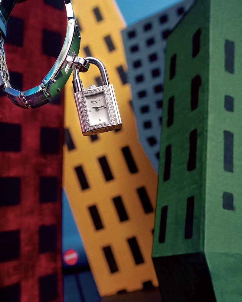 Silver wristwatch with a padlock detail hanging in front of colourful geometric buildings, blue sky above, artistic fashion photography.