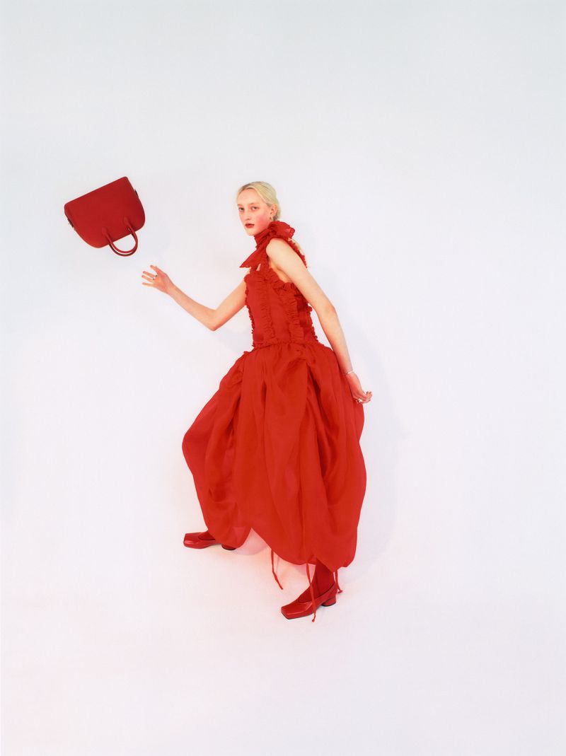 Woman in a red ruffled dress and shoes throwing a red handbag up in the air in a studio setting, fashion editorial photography.
