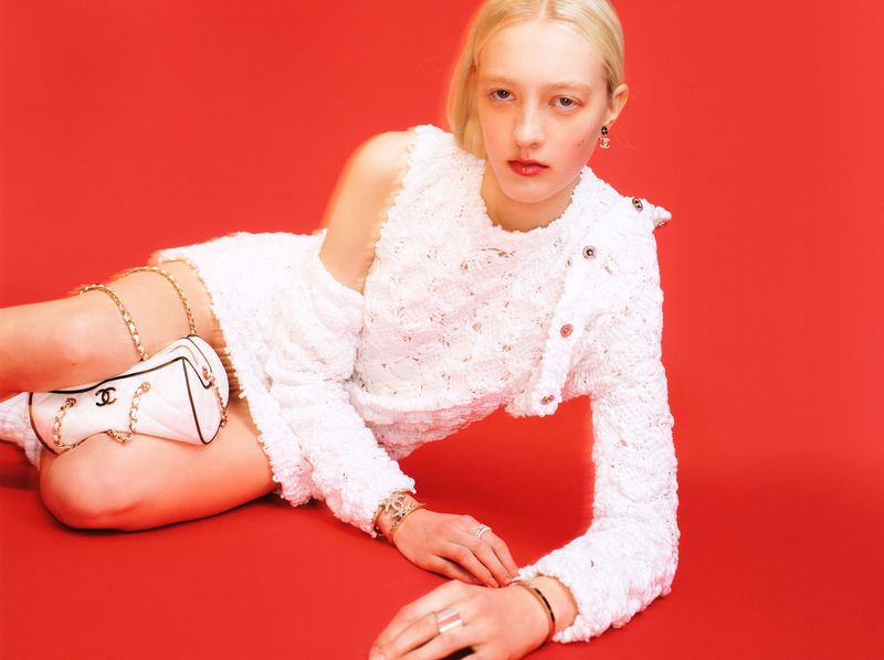 A blonde woman in a white crochet dress poses with a small white purse against a red background in a fashion photography shot.