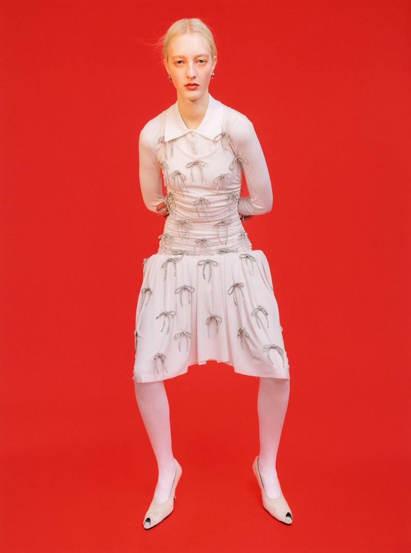 Fashion model in a white dress with bow details, sheer top, and white tights poses against a vibrant red studio backdrop, full shot.