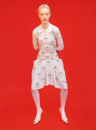 Fashion model in a white dress with bow details, sheer top, and white tights poses against a vibrant red studio backdrop, full shot.