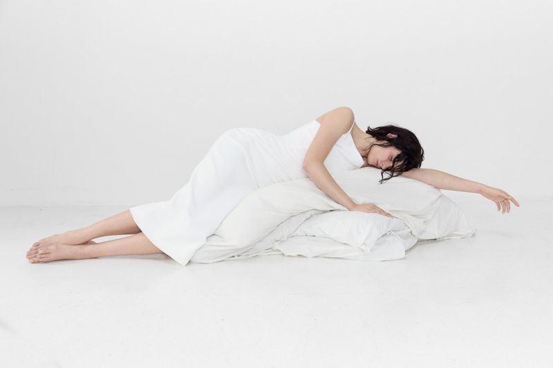 Adult woman with dark hair reclines on white pillows against a white backdrop, wearing a white dress in a brightly-lit photo studio.