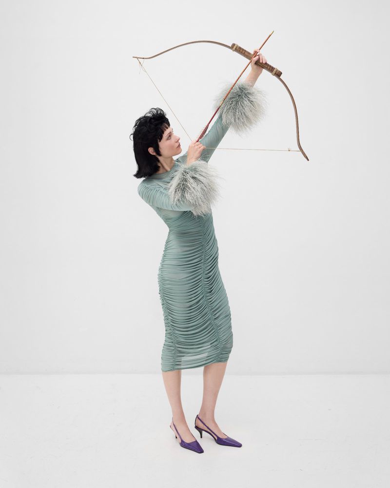 Woman in a ruched dress with fur cuffs poses in a white studio with an archery bow, looking off to the left while in purple slingback heels.