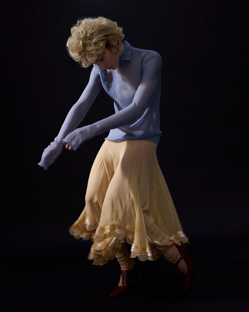 Fashionable person in motion wearing a light blue shirt and flowing yellow skirt against a dark backdrop in an indoor setting.