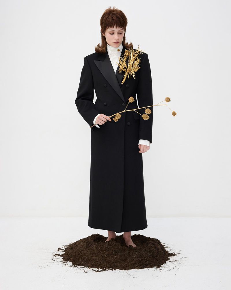 Elegant woman in a double-breasted black coat stands barefoot on soil holding a sprig with flowers against a white backdrop.