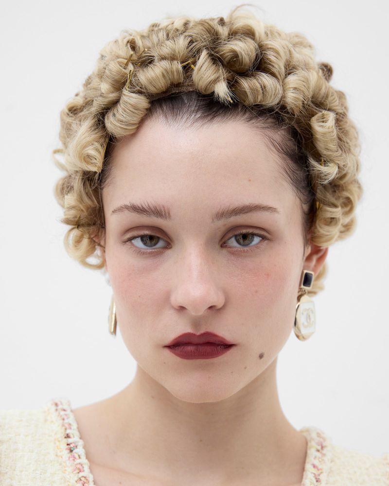 Close-up studio portrait of a blonde adult woman with curly hair, wearing statement earrings, and light yellow top against a white backdrop.