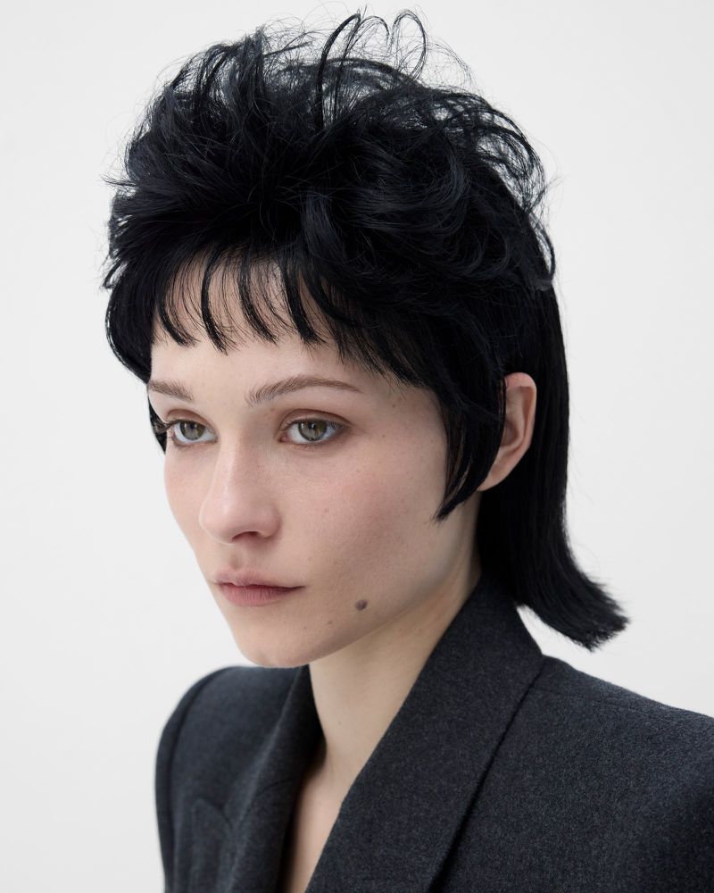Close-up studio portrait of a young adult female with black mullet hairstyle and grey blazer against a white backdrop.