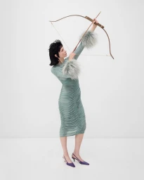 Model in a pale green ruched dress aiming a wooden bow and arrow upwards against a white studio background.