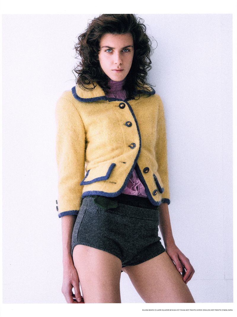 A woman poses in a yellow jacket and gray shorts against a white wall, fashion editorial style, showcasing a blend of textures and colors.