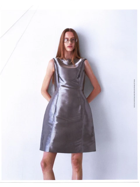 Woman with long red hair poses in a silver dress against a white backdrop. Fashion shoot with clean lines and a minimalist aesthetic.