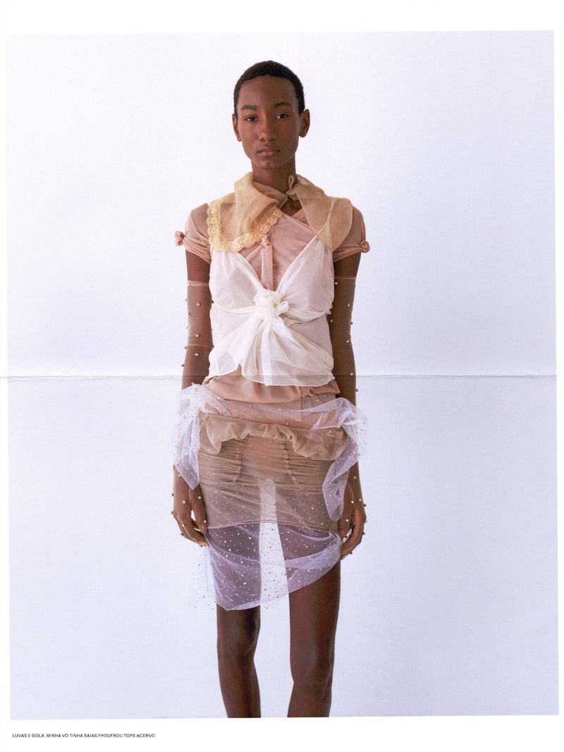 Fashion editorial shot of a Black female model wearing a sheer dress with embellishments in front of a light neutral backdrop.