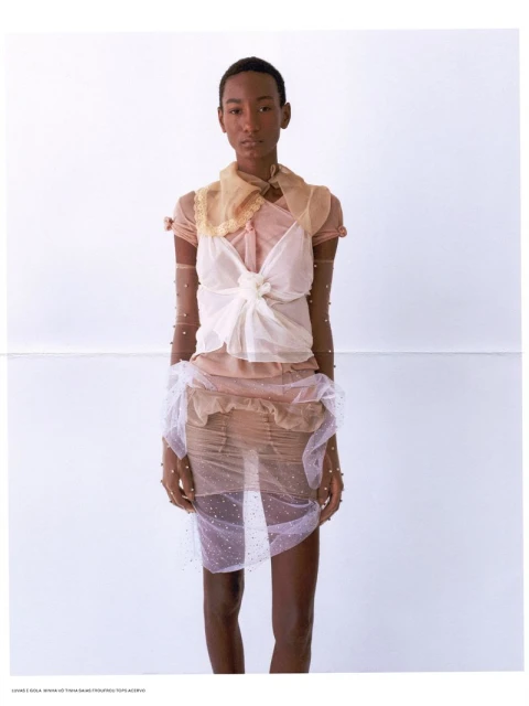 Fashion editorial shot of a Black female model wearing a sheer dress with embellishments in front of a light neutral backdrop.