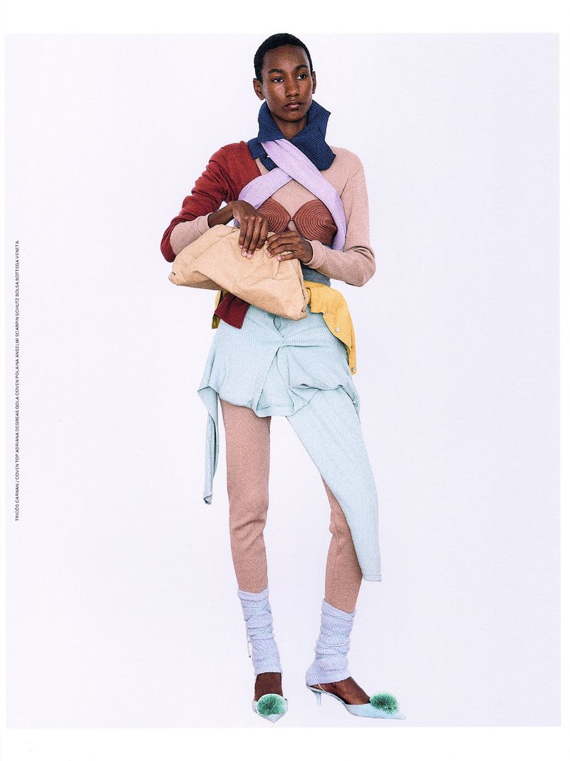 A slender, dark-skinned woman poses in a full shot, wearing a layered pastel outfit and holding a beige clutch, against a plain white background.