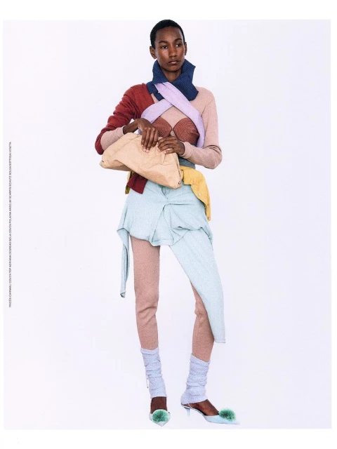 A slender, dark-skinned woman poses in a full shot, wearing a layered pastel outfit and holding a beige clutch, against a plain white background.